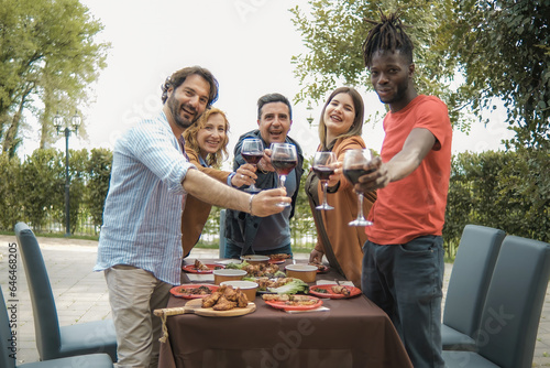 Multiethnic friends in a garden courtyard  raising wine glasses to toast directly with the viewer. Standing and smiling  they share the warmth of the evening.
