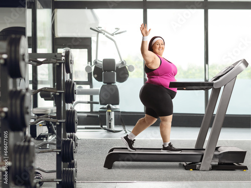 Smiling young corpulent woman walking on a treadmill at the gym and waving