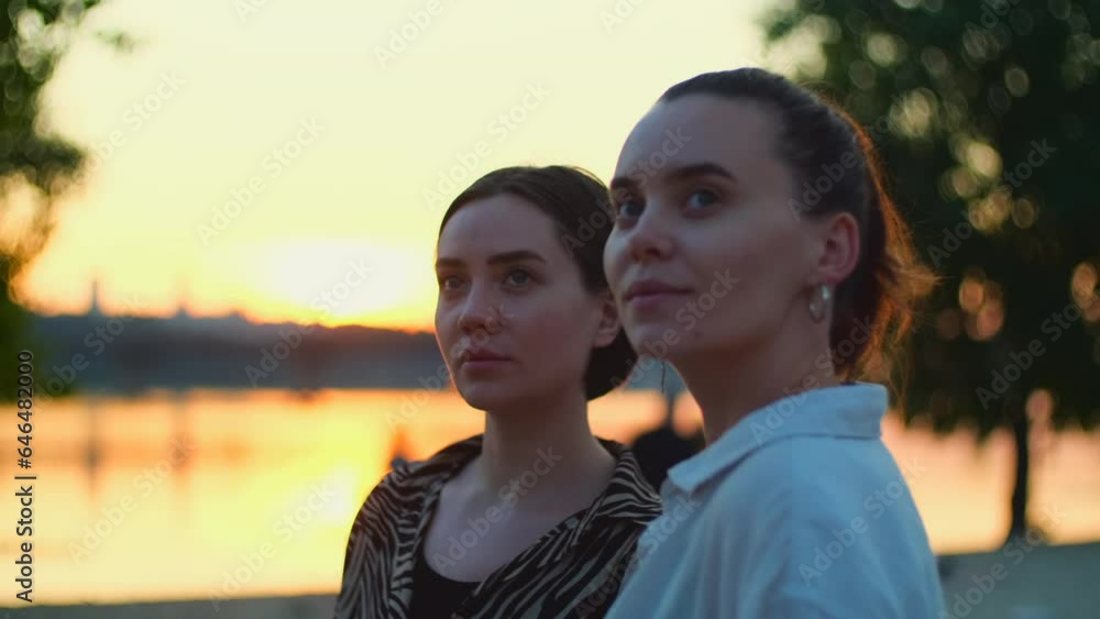 Two women enjoying sunset on the river beach. Beautiful happy girls watching sundown, carefree friends relaxing together Two women enjoying sunset on the river beach. Beautiful happy girls watching sundown, carefree friends relaxing together