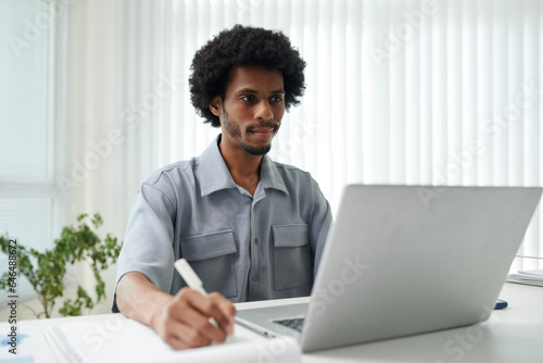 Smiling businessman reading report on laptop screen and taking notes on paper sheet