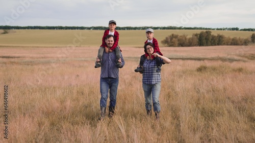 Happy mother and father walk with little boys on shoulders along in farm field