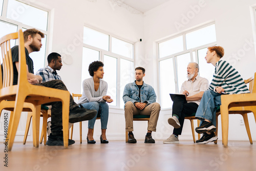 Low-angle view of confident mature male coach psychologist talking sitting in circle during group therapy with diverse and different ages people. Concept of mental health, psychotherapy, support.