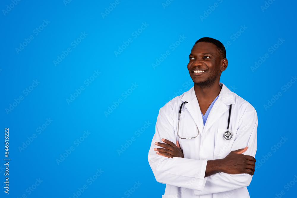 Smiling young african american guy doctor in white coat with crossed arms on chest looks at copy space Smiling young african american guy doctor in white coat with crossed arms on chest looks at copy space