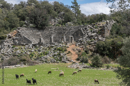 An antique panoramic landscape. A wide juicy green pasture with sheep. Ancient white stone ruins, damaged amphitheater and many small rocks are around. 