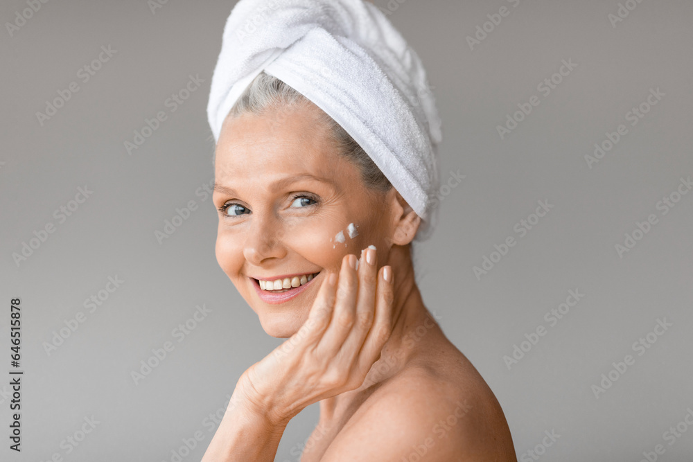 Beautiful caucasian mature woman applying moisturizing cream on face posing with towel on head and smiling at camera Beautiful caucasian mature woman applying moisturizing cream on face posing with towel on head and smiling at camera