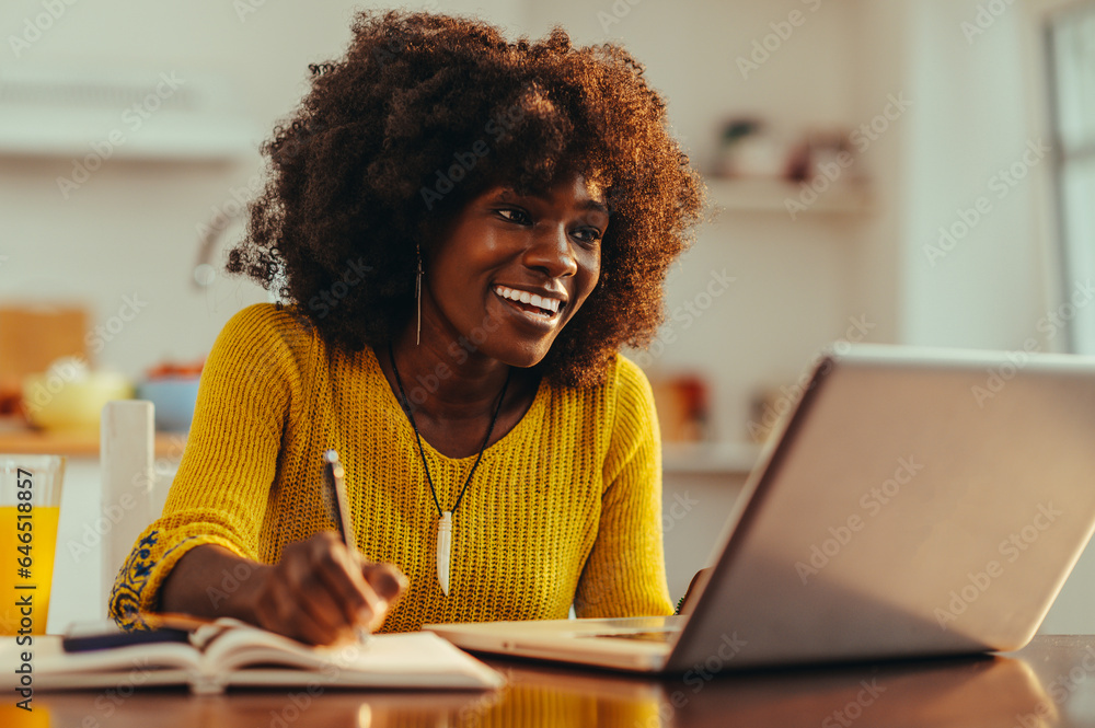 © Zamrznuti tonovi - African American woman is smiling at the laptop and writing down in the textbook. © Zamrznuti tonovi - African American woman is smiling at the laptop and writing down in the textbook.