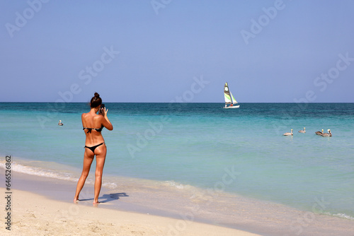 Photography Tanned slim girl in bikini taking pictures on smartphone camera standing on a tropical sandy beach
