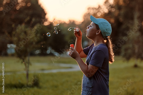 A child of 10-12 years old blows soap bubbles outside. A school-age girl enthusiastically plays in the summer park at sunset.
