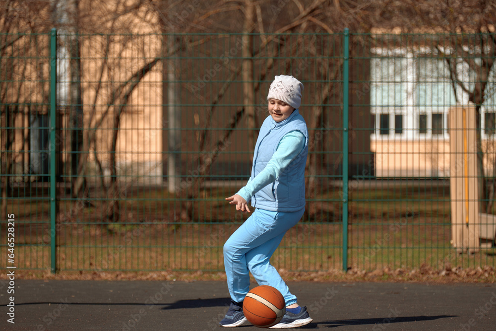 A 10-year-old girl plays basketball on an open sports ground. Child playing with a ball outdoors on an autumn day.