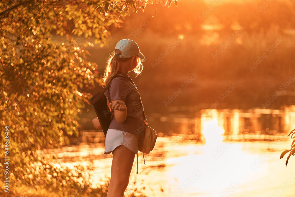 A teenage girl 12-13 years old stands on the banks of the river and enjoys the beauty of nature at sunset on a summer day. A teenage girl 12-13 years old stands on the banks of the river and enjoys the beauty of nature at sunset on a summer day.