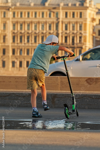 A boy of 8-10 years old rides a two-wheeled scooter through the puddles in the summer.