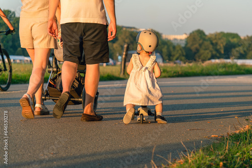 Happy family outdoors at sunset. A little girl in a white dress and helmet rides along the path on a blue scooter, and her parents walk nearby. Green fields and trees in the background