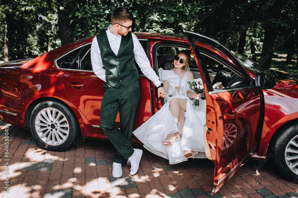 A stylish groom in a suit and a beautiful bride in sunglasses are hugging near a red car. Wedding photography, portrait, lifestyle.