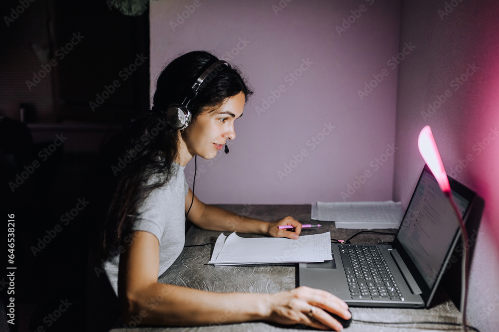 A young girl, a woman works, sitting on a laptop in headphones with a microphone at night, sitting at a table. Photography, portrait, lifestyle. A young girl, a woman works, sitting on a laptop in headphones with a microphone at night, sitting at a table. Photography, portrait, lifestyle.