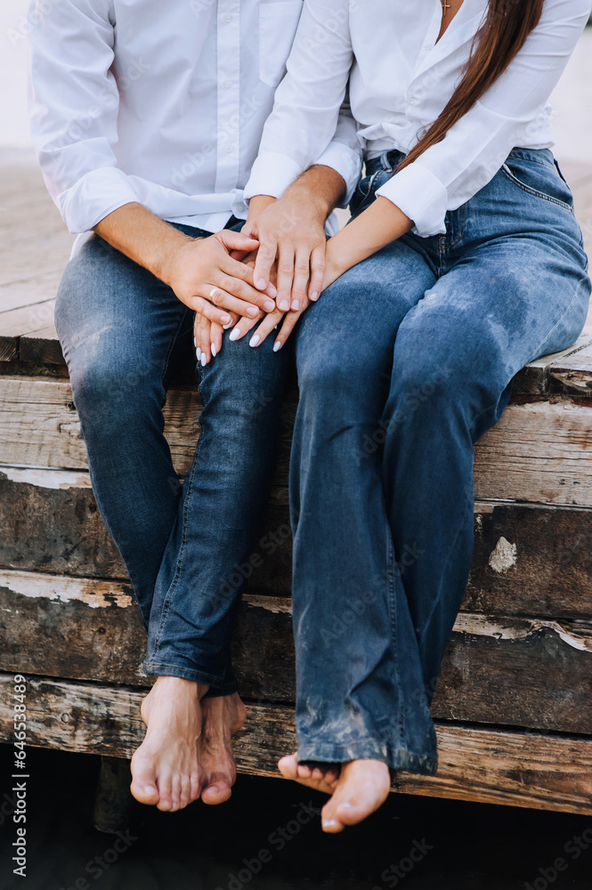 Barefoot man and woman in white shirts, blue wet jeans sitting on a wooden pier near the sea in summer. Close-up photography, portrait, vacation lifestyle. Barefoot man and woman in white shirts, blue wet jeans sitting on a wooden pier near the sea in summer. Close-up photography, portrait, vacation lifestyle.