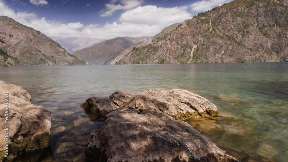 Lake in mountain gorge timelapse. Shooting near shore, next to large boulder. Small ripples from wind on surface of water. Sky is reflected in water. Calmness and balance concept. Cinematic video Lake in mountain gorge timelapse. Shooting near shore, next to large boulder. Small ripples from wind on surface of water. Sky is reflected in water. Calmness and balance concept. Cinematic video