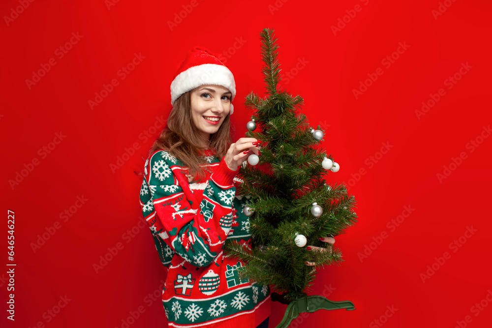 young cute girl in Christmas sweater and santa hat decorates the Christmas tree on red background young cute girl in Christmas sweater and santa hat decorates the Christmas tree on red background