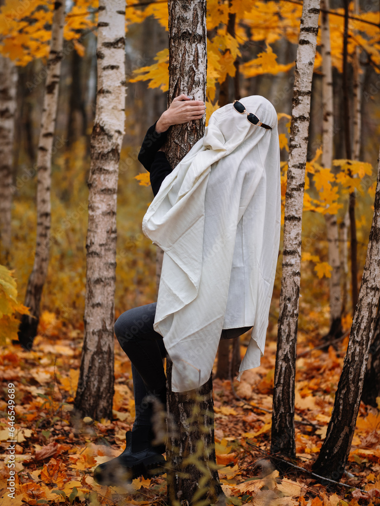 A funny girl in a Halloween ghost costume wearing sunglasses climbs a tree. Festive costume made from a sheet A funny girl in a Halloween ghost costume wearing sunglasses climbs a tree. Festive costume made from a sheet