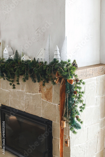 Modern christmas trees, houses, pine cones and spruce branches on fireplace mantel with brass bells and ribbon. Stylish decorated christmas fireplace in minimal scandinavian living room