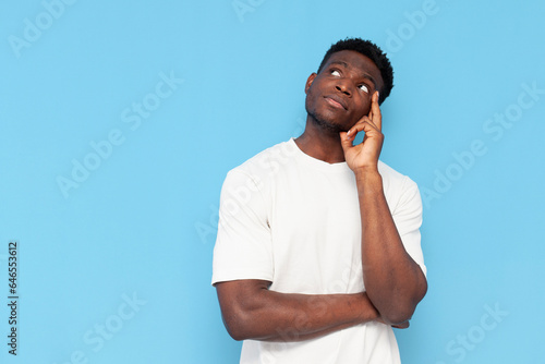 pensive guy african american in white t-shirt plans and thinks on blue isolated background, man dreams and imagines