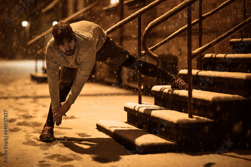 Young man stretching while out jogging and exercising at night during winter and snow in the city