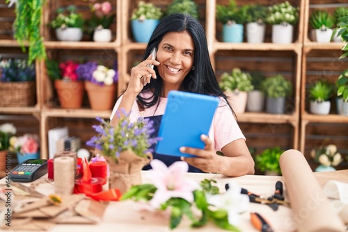 Middle age hispanic woman florist talking on smartphone using touchpad at florist