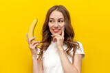 young pensive girl holding banana and smiling on yellow isolated background, woman dreams and sexualizes the fruit