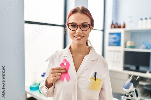 Young caucasian woman scientist smiling confident holding pink cancer ribbon at laboratory