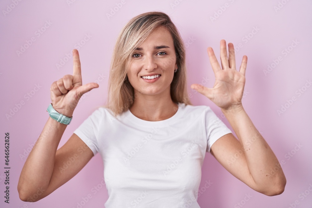Young blonde woman standing over pink background showing and pointing up with fingers number seven while smiling confident and happy. Young blonde woman standing over pink background showing and pointing up with fingers number seven while smiling confident and happy.