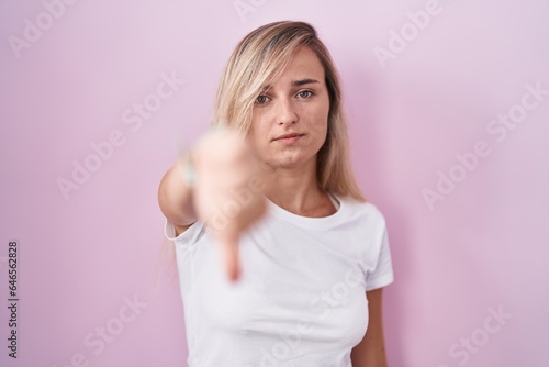 Young blonde woman standing over pink background looking unhappy and angry showing rejection and negative with thumbs down gesture. bad expression.
