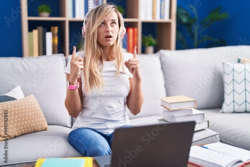 Young blonde woman studying using computer laptop at home amazed and surprised looking up and pointing with fingers and raised arms.