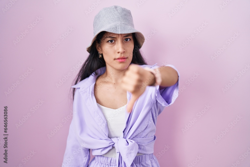 Young hispanic woman standing over pink background wearing hat looking unhappy and angry showing rejection and negative with thumbs down gesture. bad expression. Young hispanic woman standing over pink background wearing hat looking unhappy and angry showing rejection and negative with thumbs down gesture. bad expression.