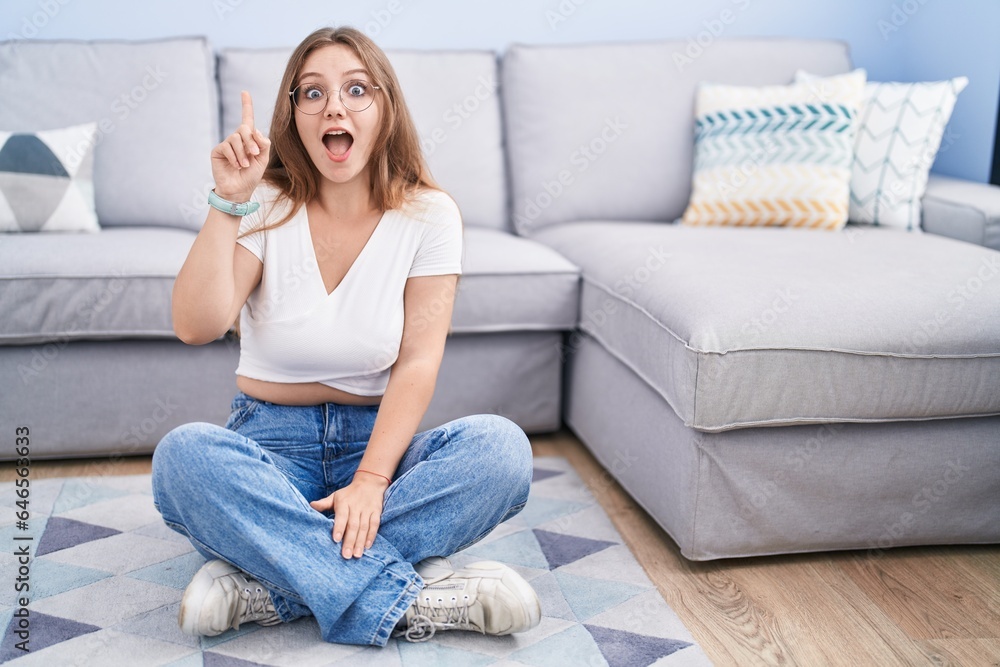 Young caucasian woman sitting on the floor at the living room pointing finger up with successful idea. exited and happy. number one. Young caucasian woman sitting on the floor at the living room pointing finger up with successful idea. exited and happy. number one.