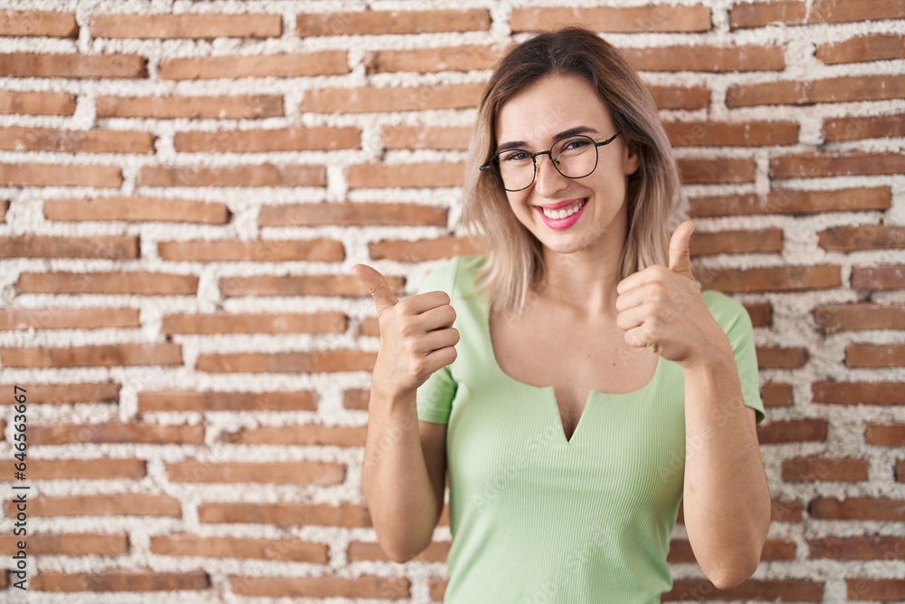 Young beautiful woman standing over bricks wall success sign doing positive gesture with hand, thumbs up smiling and happy. cheerful expression and winner gesture. Young beautiful woman standing over bricks wall success sign doing positive gesture with hand, thumbs up smiling and happy. cheerful expression and winner gesture.