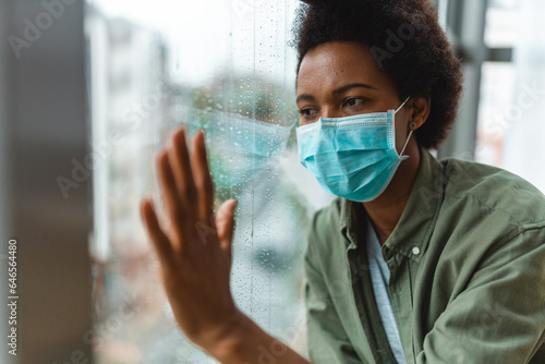 Devastated young female holding hand on window in hospital and looking hopeless into the distance wearing protective face mask. Sad African American woman looking through window on rainy day.