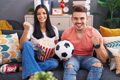 Young hispanic couple football hooligans holding ball and eating popcorn screaming proud, celebrating victory and success very excited with raised arm