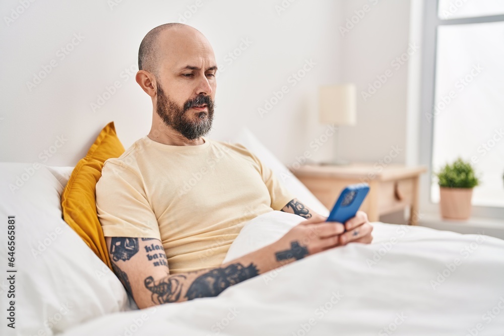 Young bald man using smartphone sitting on bed at bedroom Young bald man using smartphone sitting on bed at bedroom
