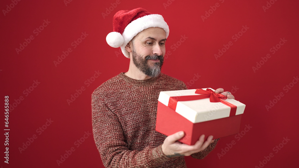 Young bald man wearing christmas hat unpacking gift over isolated red background Young bald man wearing christmas hat unpacking gift over isolated red background