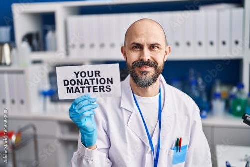 Young hispanic man working at scientist laboratory holding you donation matters banner looking positive and happy standing and smiling with a confident smile showing teeth