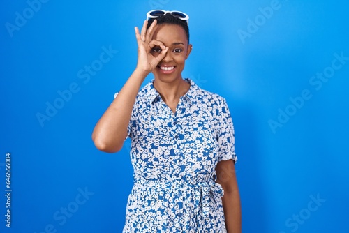 African american woman standing over blue background doing ok gesture with hand smiling, eye looking through fingers with happy face.