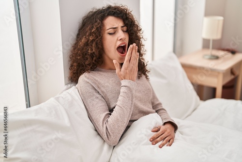 Young hispanic woman sitting on bed yawning at bedroom photo