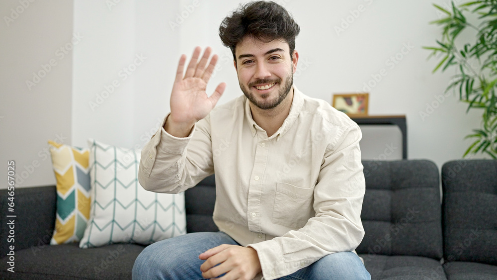 Young hispanic man saying hello with hand sitting on sofa at home Young hispanic man saying hello with hand sitting on sofa at home
