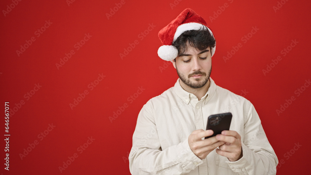 Young hispanic man wearing christmas hat using smartphone over isolated red background Young hispanic man wearing christmas hat using smartphone over isolated red background