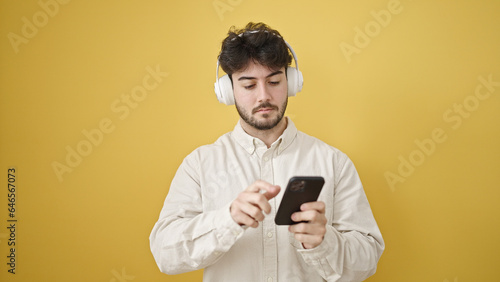 Young hispanic man using smartphone wearing headphones over isolated yellow background