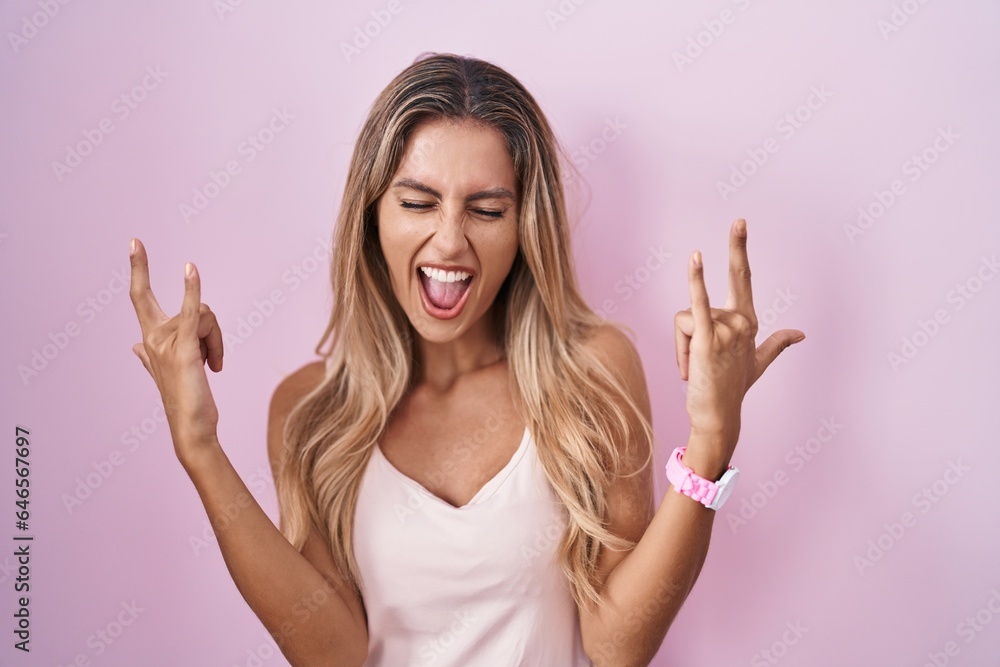 Young blonde woman standing over pink background shouting with crazy expression doing rock symbol with hands up. music star. heavy music concept. Young blonde woman standing over pink background shouting with crazy expression doing rock symbol with hands up. music star. heavy music concept.