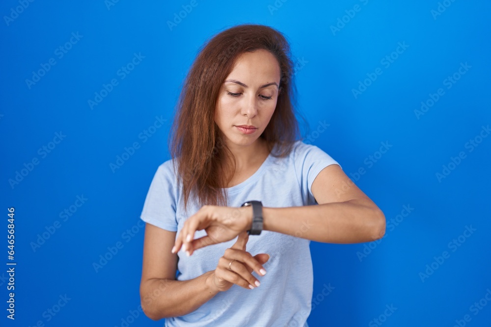 Brunette woman standing over blue background checking the time on wrist watch, relaxed and confident Brunette woman standing over blue background checking the time on wrist watch, relaxed and confident