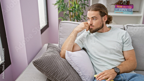 Young hispanic man sitting on sofa holding smartphone thinking at home