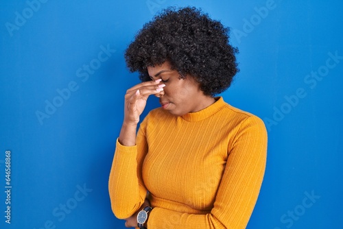 Black woman with curly hair standing over blue background tired rubbing nose and eyes feeling fatigue and headache. stress and frustration concept.
