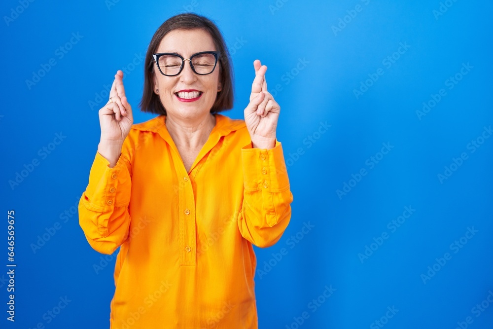 Middle age hispanic woman wearing glasses standing over blue background gesturing finger crossed smiling with hope and eyes closed. luck and superstitious concept. Middle age hispanic woman wearing glasses standing over blue background gesturing finger crossed smiling with hope and eyes closed. luck and superstitious concept.