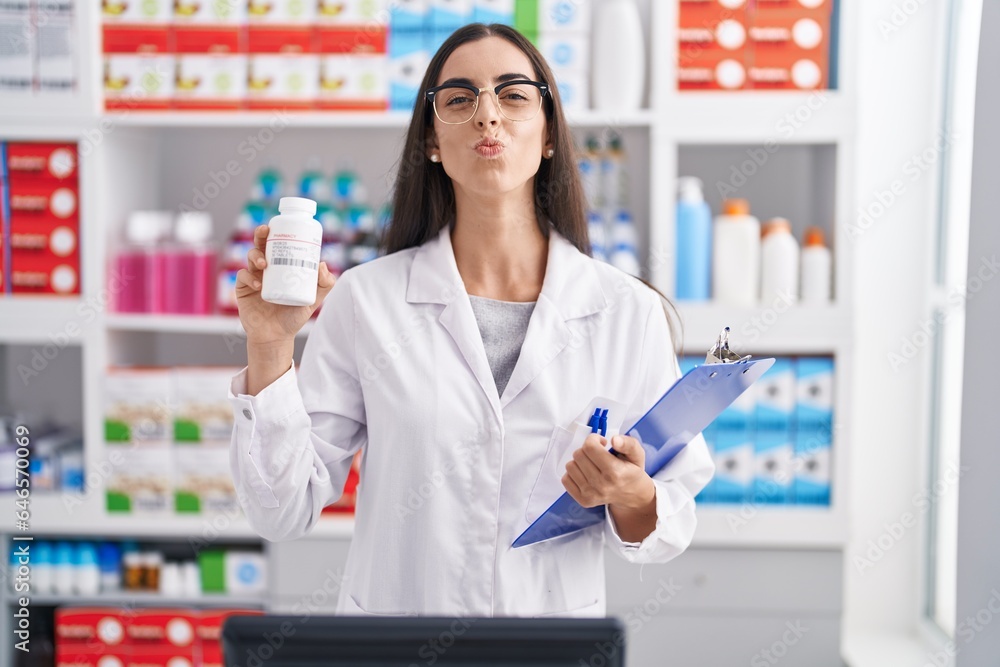 Young brunette woman working at pharmacy drugstore holding pills looking at the camera blowing a kiss being lovely and sexy. love expression.